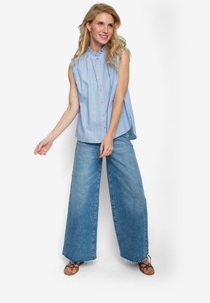 Blauw-witte gestreepte mouwloze blouse met een gerimpelde kraag en knopen aan de voorkant, gecombineerd met lichtblauwe wijde denim jeans. Bruine sandalen met bandjes.