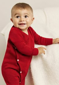 Punainen neulotut haalari, jossa on V-pääntie, pitkät hihat ja napit sivussa. Lapsi hymyilee nojaten vaaleaan pintaan.