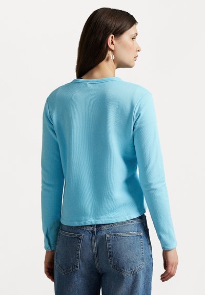 Himmelblaues Langarmoberteil mit gerippter Textur, Rundhalsausschnitt und enganliegenden Ärmeln. Kombiniert mit hochtaillierten blauen Jeans.