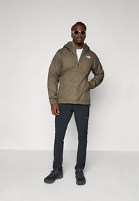 Olivovo zelená bunda na zips s kapucňou, s bielym logom, v páre s tmavými nohavicami a čiernymi turistickými topánkami. Hladká textúra, voľný strih.