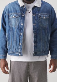 Man som bär en blå denimjacka med vit shearlingkrage, en vit t-shirt med tryck och svarta och vita vertikalt randiga byxor.