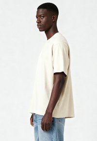 Bēša kokvilnas T-krekls ar īsām piedurknēm un brīvu piegriezumu, valkāts kopā ar gaiši ziliem džinsiem. Viens tonis un neformāls dizains.