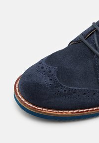 Chaussure en daim bleu marine avec des perforations au niveau de la toe, des détails en cuir, et une semelle en caoutchouc bleue contrastante. Forme arrondie avec un système de laçage.