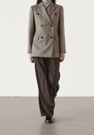 Femme portant un blazer croisé taupe sur un col roulé gris clair et un pantalon ample marron foncé avec des chaussures noires, marchant vers l'avant.