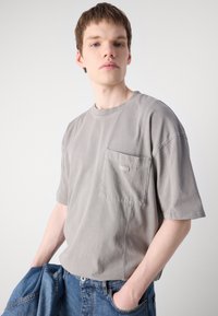 Grå bomulds T-shirt med korte ærmer, en lomme på venstre bryst og synlige syninger. Sammen med denim jeans.