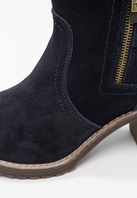 Bottine en daim bleu marine avec une finition texturée, dotée d'une fermeture éclair dorée et de détails cousus. Talon bas et bout arrondi.