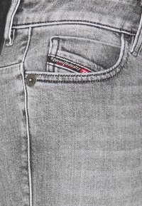 Jeans in denim grigio con tasca frontale dotata di un'etichetta logo rossa e nera, chiusura con bottone metallico e cuciture in contrasto. Tessuto testurizzato.