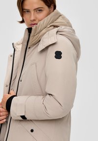 Beige gewatteerde jas met hoge kraag, zwarte hardware, contrasterende manchetten en een logo op de schouder. Zachte textuur en gewatteerd ontwerp.