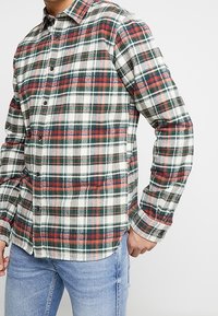 Karo-Flanellhemd in Weiß-, Grün- und Rottönen mit Knopfleiste, spitzem Kragen und langen Ärmeln, getragen über blauen Jeans.