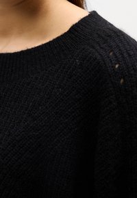 Gros plan d'une personne portant un pull en maille noir avec un col rond côtelé et un motif de point texturé.