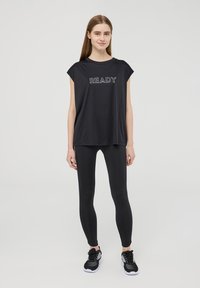 T-shirt atletico nero con la scritta "READY" in lettere bianche, abbinato a leggings neri e scarpe da ginnastica nere. Tessuto liscio, vestibilità comoda.