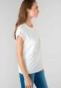 Top bianco a maniche corte con due tasche frontali, scollo rotondo e delicati dettagli in rilievo sulle maniche. Indossato con jeans blu.