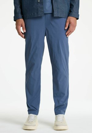 Homme portant un pantalon décontracté bleu, une veste marine et des baskets beige, debout devant un fond blanc uni.