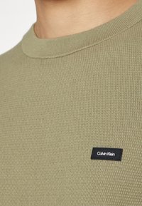 Gros plan sur la partie supérieure de la poitrine d'une personne portant un pull texturé vert olive avec une petite étiquette noire du logo Calvin Klein.