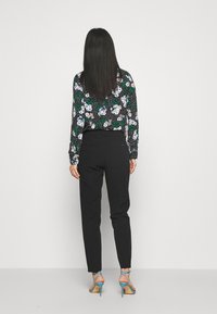 Pantalons noirs et un haut à manches longues à motifs floraux avec des accentements verts et blancs. Le modèle porte des sandales à talons bleues, vu de dos.