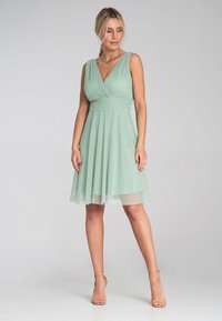 Kurzes grünes Kleid mit taillierter Blende, V-Ausschnitt und geschichteter Rock. Hergestellt aus leichtem Stoff mit fließender Textur.