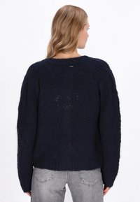 Sötétkék kötött pulóver texturált mintával, széles ujjakkal és bordázott aljjal. A hátsó nyakkivágásnál egy diszkrét logó címke található.