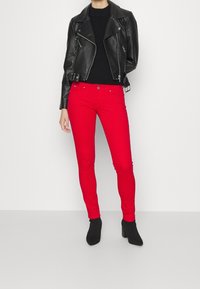 Jeans skinny rojos combinados con una chaqueta de moto de cuero negra y un suéter negro ajustado. La modelo lleva botas de tobillo negras. Textura suave.