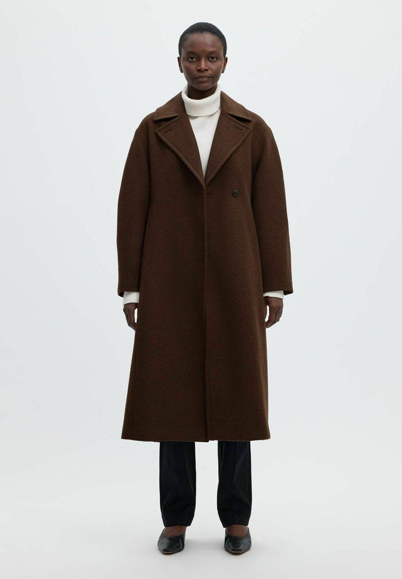 Femme debout portant un long manteau marron foncé par-dessus un col roulé blanc, un pantalon noir et des chaussures noires sur un fond clair uni.