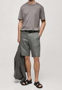 T-shirt gris à manches courtes, shorts gris clair, ceinture noire et sandales noires ; veste posée sur le bras ; tissu lisse, design simple.
