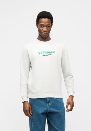 Junger Mann, der ein hellgraues Sweatshirt mit Rundhalsausschnitt und dem Aufdruck "T.W.Y Jeans" sowie eine blaue Jeans trägt, vor einem schlichten weißen Hintergrund steht.