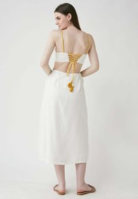 Weißes rückenfreies Oberteil mit gelben Schnürdetails und Quastenakzenten, kombiniert mit einem weißen Midikleid. Modell trägt neutrale Sandalen.