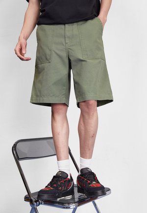 Mand iført olivengrønne shorts, hvide sokker og sorte sneakers står på en sort klapstol mod en grå baggrund.