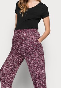 Woman wearing black short-sleeve top and high-waisted maroon pants with white and black floral print, hand in pocket, against plain background.