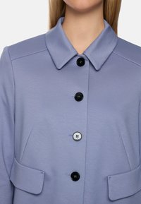 Lilac coat featuring a collared neckline, three black buttons, one white button, and topstitched details. Includes two front pockets.