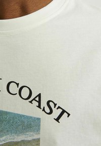 Krémové bavlnené tričko s fotografickou potlačou plážovej scény a textom "COAST" v tučných čiernych písmenách nad ňou.