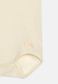 Bodysuit pour bébé beige clair à manches courtes, avec une texture douce et une fermeture à pression au bas. Étiquette "Sanetta" en orange clair cousue sur le côté.