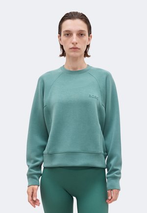 Γυναίκα με κοντά βρεγμένα μαλλιά φοράει ένα απλό τιρκουάζ φούτερ με μικρό λογότυπο «BORG» και ταιριαστό τιρκουάζ κολάν, στέκεται μπροστά σε ανοιχτό φόντο.