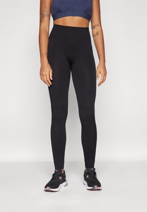 Leggings noirs côtelés avec une taille haute, fabriqués dans un matériau extensible. Portés avec des chaussures de sport noires et roses. Peau visible au niveau du ventre.