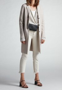 Manteau long beige clair en tissu texturé, superposé à une chemise gris clair, associé à un jean blanc court et des sandales à talons noires à lanières.