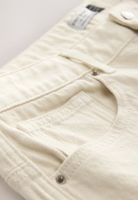 Pantalon beige en coton avec fermeture éclair, bouton et une poche avant. Le tissu présente un motif texturé subtil.
