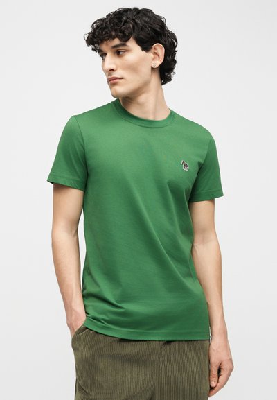 Νέος άνδρας με σγουρά μαλλιά που φοράει πράσινο κοντομάνικο μπλουζάκι με μικρό λογότυπο ζέβρας στο στήθος και χακί παντελόνι κοτλέ, κοιτάζοντας προς τα αριστερά.