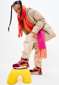 Sciarpa rosa e arancione, giacca puffer beige, pantaloni beige e sneakers multimateriale bordeaux con accenti beige, in piedi su uno sgabello giallo.