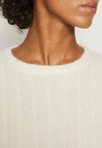 Cremefarbener Pullover mit strukturiertem Rippmuster, weiter runder Ausschnitt und weichem Stoff, der ein lässiges, aber dennoch elegantes Design zeigt.