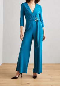 Combinaison bleu sarcelle avec un décolleté en V profond, des manches 3/4 et une ceinture à la taille avec un accent métallique circulaire. Fabriquée en tissu lisse et élastique.