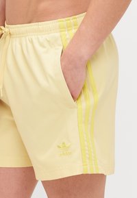 Κίτρινα αθλητικά σορτς με ελαστική μέση, πλαϊνές τσέπες και τρεις αντίθετες κίτρινες ρίγες κατά μήκος των πλευρών. Διαθέτει κεντημένο λογότυπο.