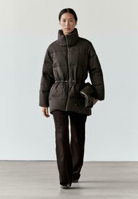 Brązowa pikowana kurtka z wysokim kołnierzem, taliowana z zamkiem błyskawicznym; w zestawieniu z ciemnymi szerokimi spodniami i małą czarną torebką.