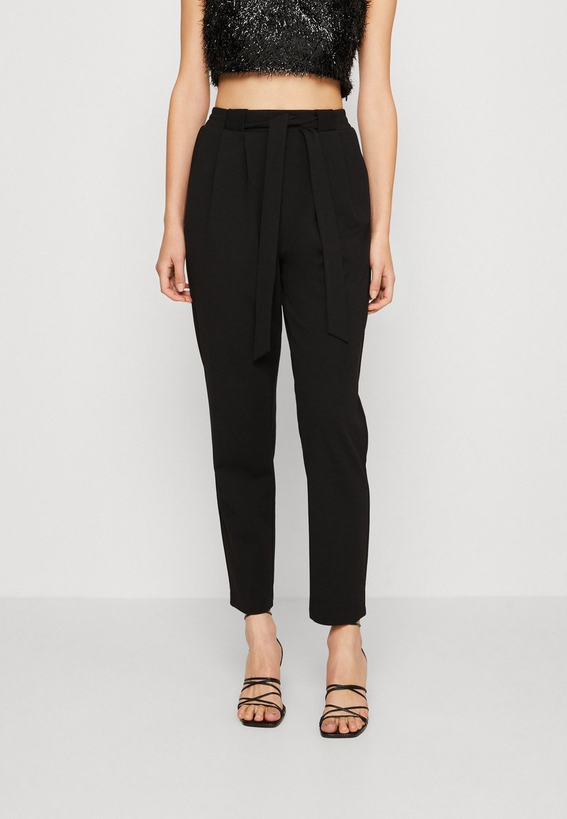 Pantalon noir taille haute avec ceinture nouée, ajusté à la taille et resserré aux chevilles, associé à des talons noirs à brides.