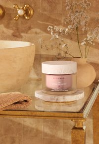 Vispad kroppskräm i en glasburk med en ljus rosa gradient och ett trälock. Inkluderar en rund marmorplatta och en beige texturerad duk i närheten.