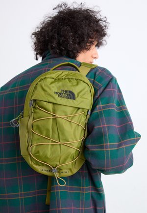 Person wearing green plaid jacket with curly hair carrying small olive green The North Face backpack with front elastic cords.