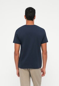 T-shirt in cotone navy con maniche corte, scollatura a girocollo e una consistenza liscia. Indossato con pantaloni chiari, girato di spalle rispetto all'osservatore.