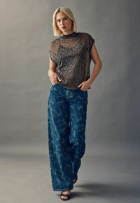 Gennemsigtig, metallisk bronze top med korte ærmer parret med brede blå denimbukser med et legende sløjfemønster. Sorte åben-tå hæle.