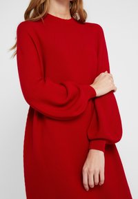 Femme portant une longue robe en maille rouge avec des manches bouffantes, debout avec un bras croisé sur l'autre devant un fond uni.