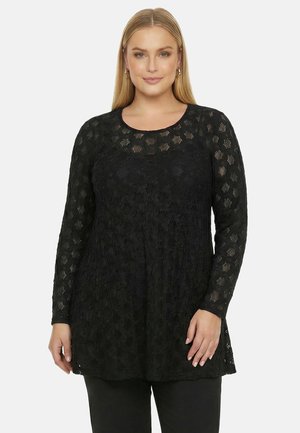 Frau mit glatten blonden Haaren, die eine lange schwarze Spitzenbluse mit langen Ärmeln und schwarze Hosen trägt, steht vor einem weißen Hintergrund.