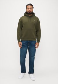 Φούτερ σε ελιά απόχρωση με μπροστινή τσέπη και κουκούλα με ρυθμιζόμενο κορδόνι, σετ με μπλε τζιν και λευκά αθλητικά παπούτσια πάνω σε ουδέτερο φόντο.