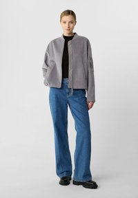 Graue Bomberjacke mit glatter Oberfläche, ausgestattet mit einem Reißverschluss und Seitentaschen. Kombiniert mit lässigen blauen Jeans und schwarzem Schuhwerk.